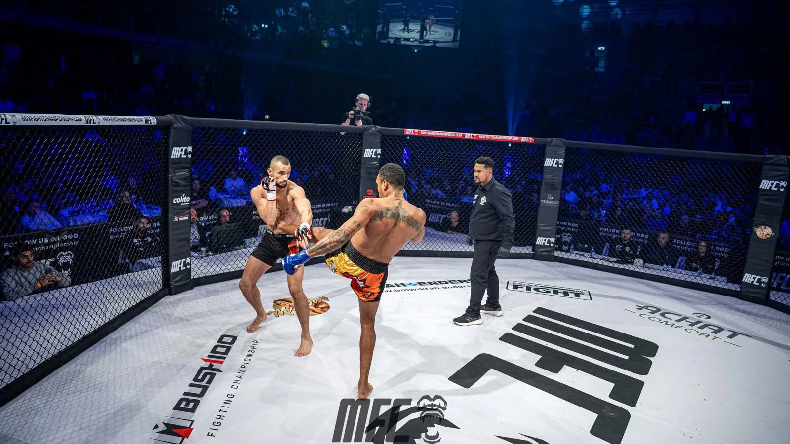 Zwei Kämpfer im Käfig der Mix Fight Championship, einer tritt zu, daneben der Ringrichter; auf dem weißen Käfigboden große Firmenlogos