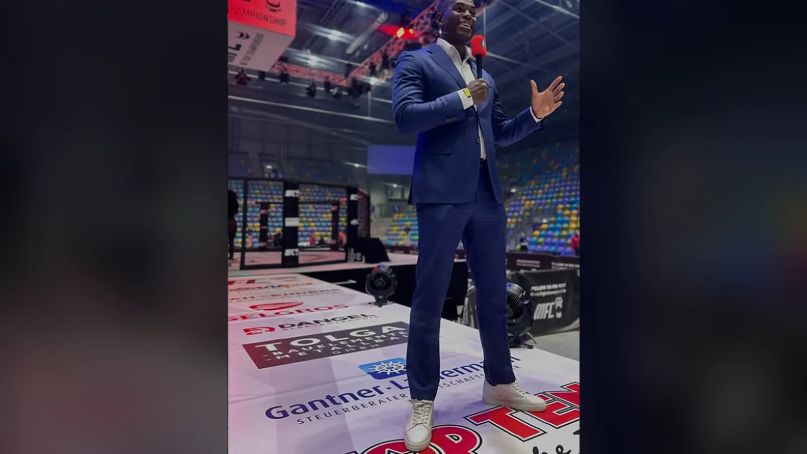Moderator im blauen Anzug spricht ins Mikrofon auf dem Laufsteg der Mix Fight Championship; auf dem Laufstegboden mehrere Firmenlogos, dahinter Käfig und Tribüne