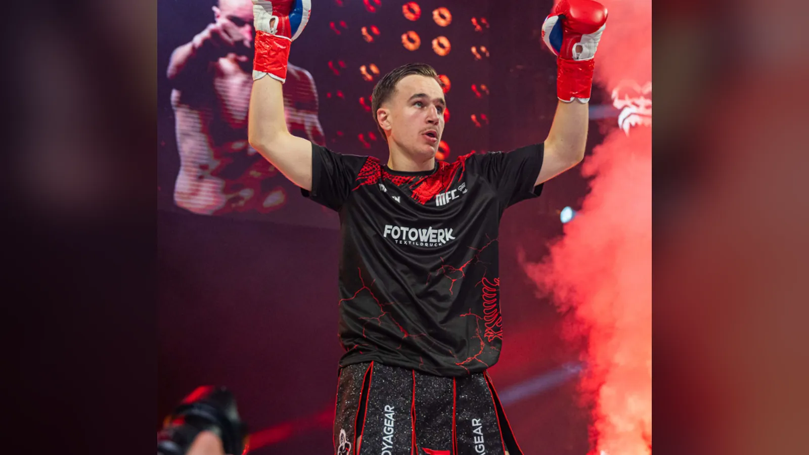 Kämpfer jubelt beim Einlauf mit erhobenen Boxhandschuhen; er trägt ein schwarz-rotes Walkout-Shirt mit dem Firmenlogo Fotowerk und dem MFC-Logo